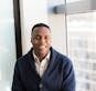 A professional black man smiling while seated by a window, embodying modern business vibes.